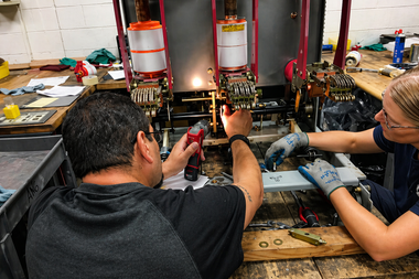 Two technicians repairing electronics on a workbench with tools and wiring.
