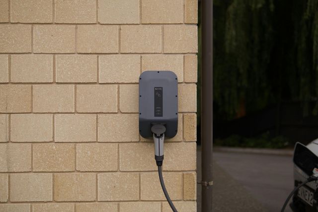 Wall-mounted electric vehicle charger on a beige brick exterior wall with a cable plugged in.