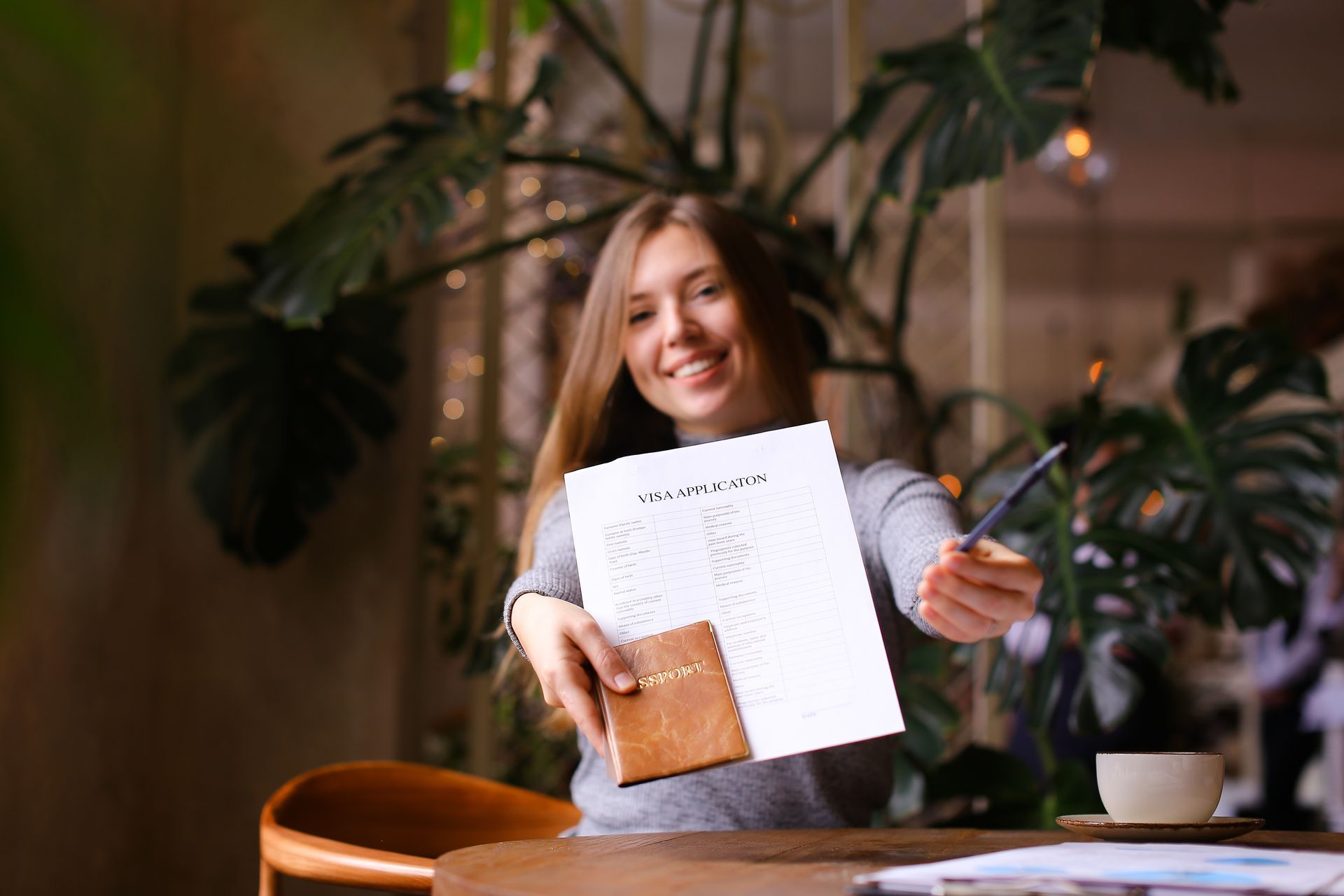 Woman smiles, holding paperwork and a pen, offering them forward near a plant and coffee.