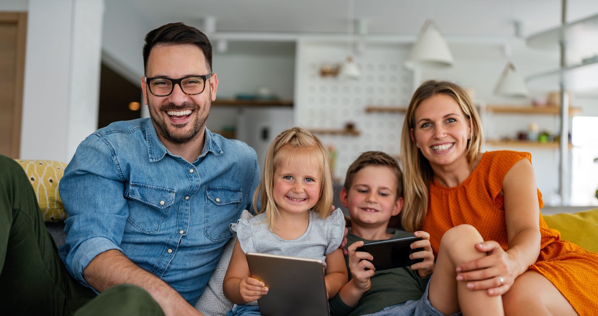 A smiling family sits on a couch in a bright living room, with the children playing on electronic devices.