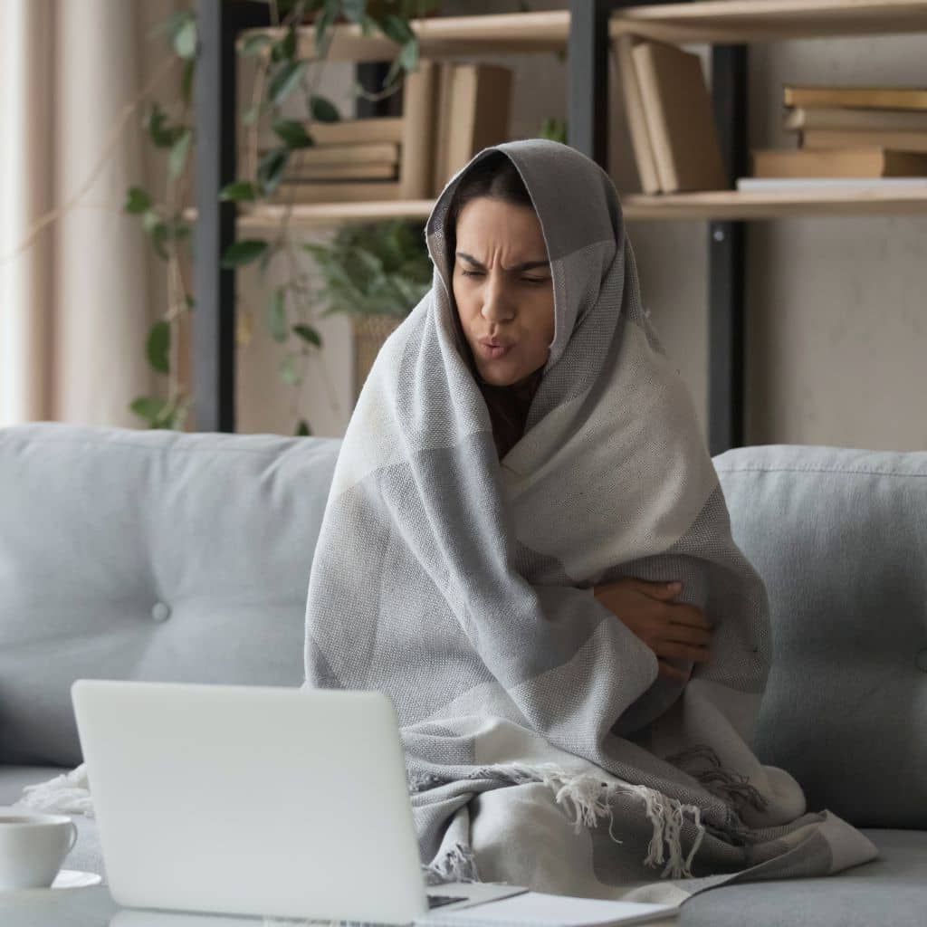 Woman wrapped in a blanket, looking cold, working on a laptop on a couch.