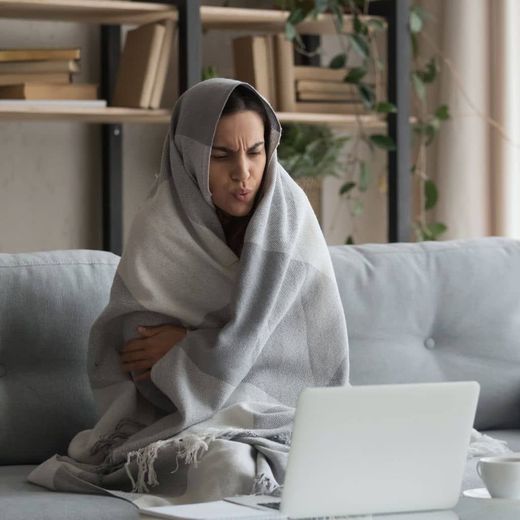 A woman wrapped in a blanket is sitting on a couch using a laptop computer.