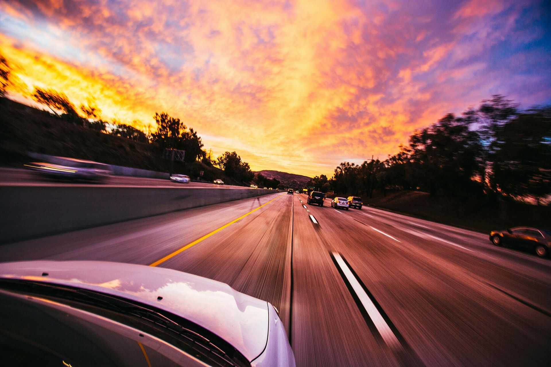 Highway at sunset, cars speeding on asphalt road with colorful sky.