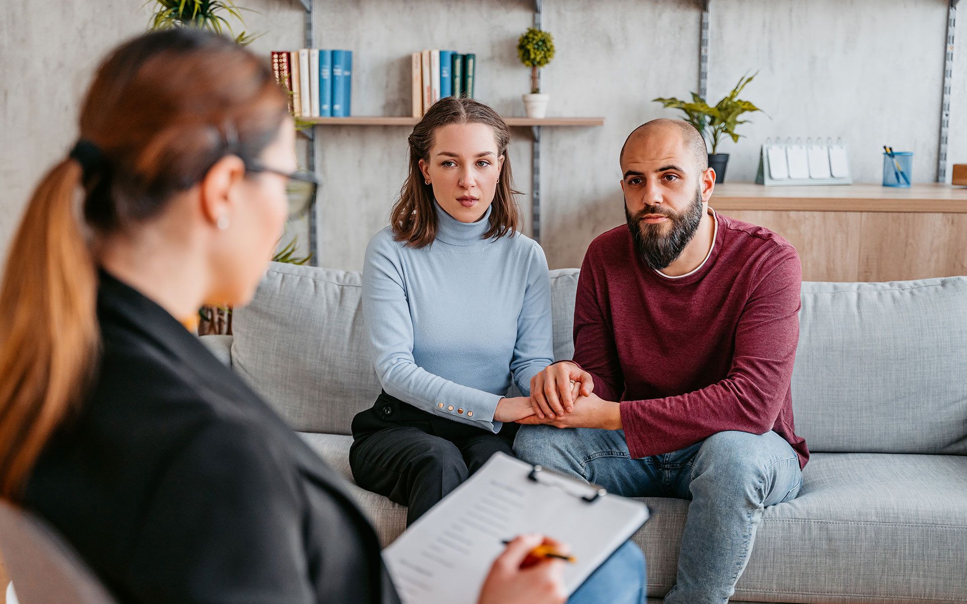 A man and a woman are sitting on a couch holding hands while a woman holds a clipboard.