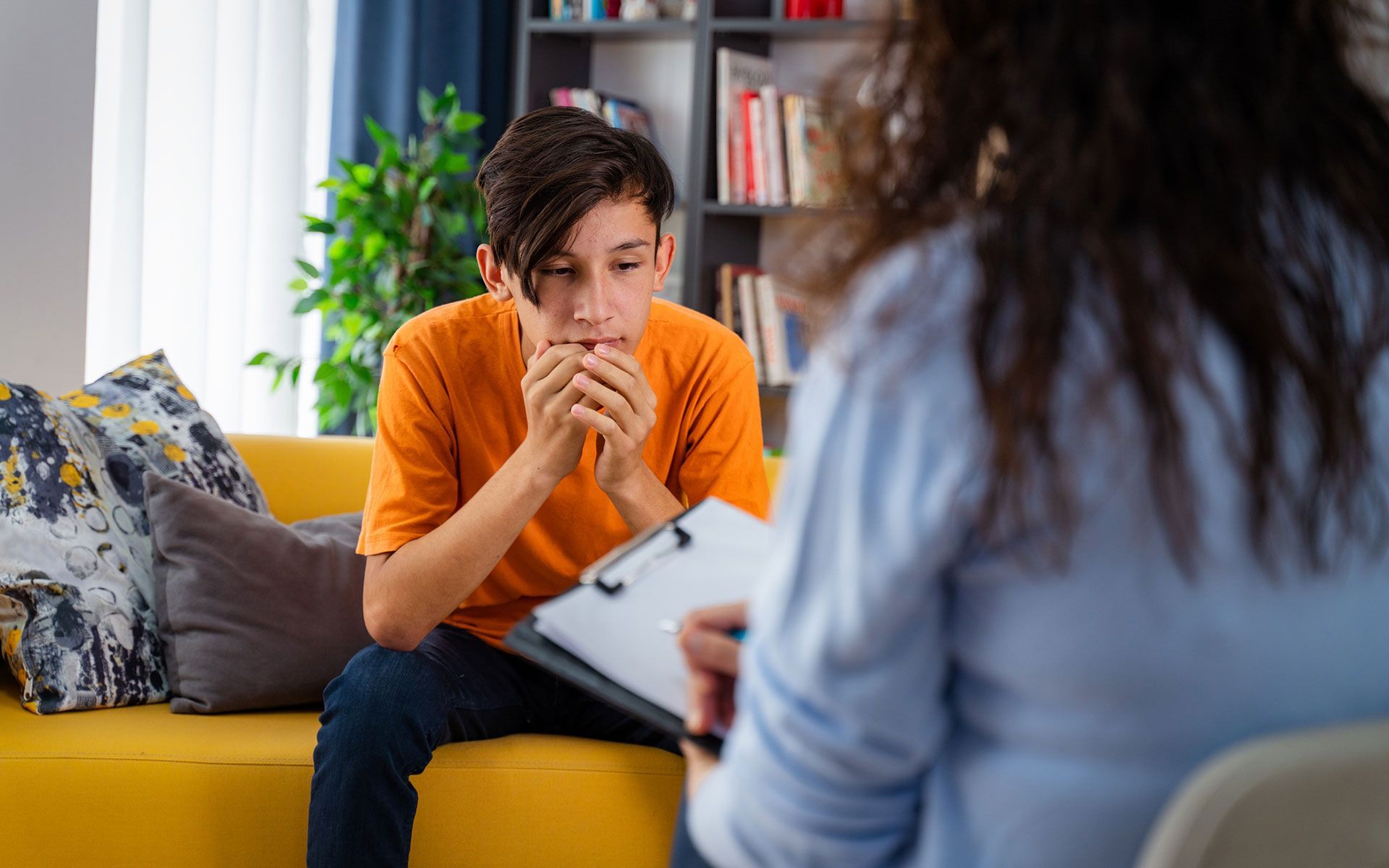 A young boy is sitting on a yellow couch talking to a woman.