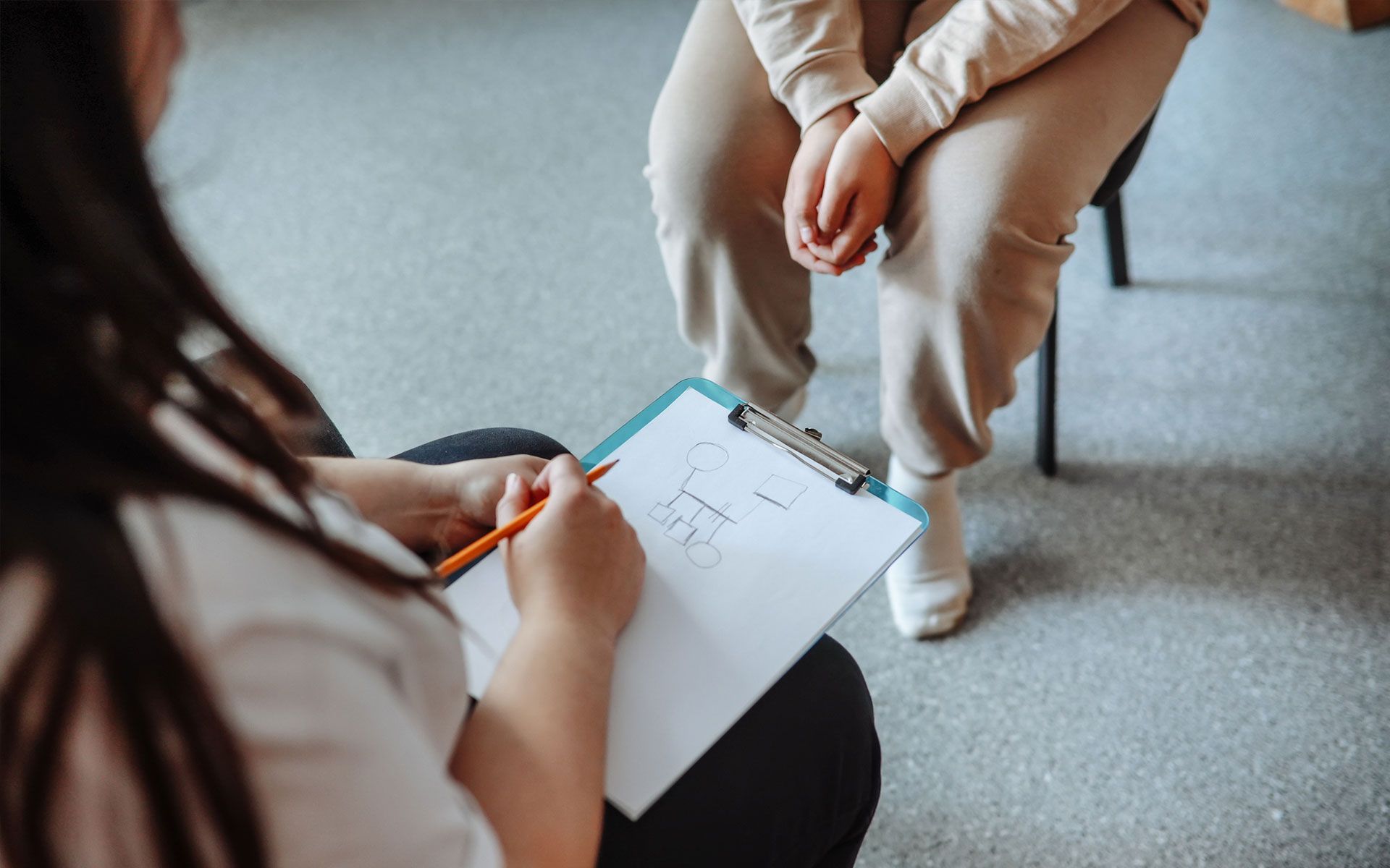 A woman is sitting in a chair talking to another woman while holding a clipboard.
