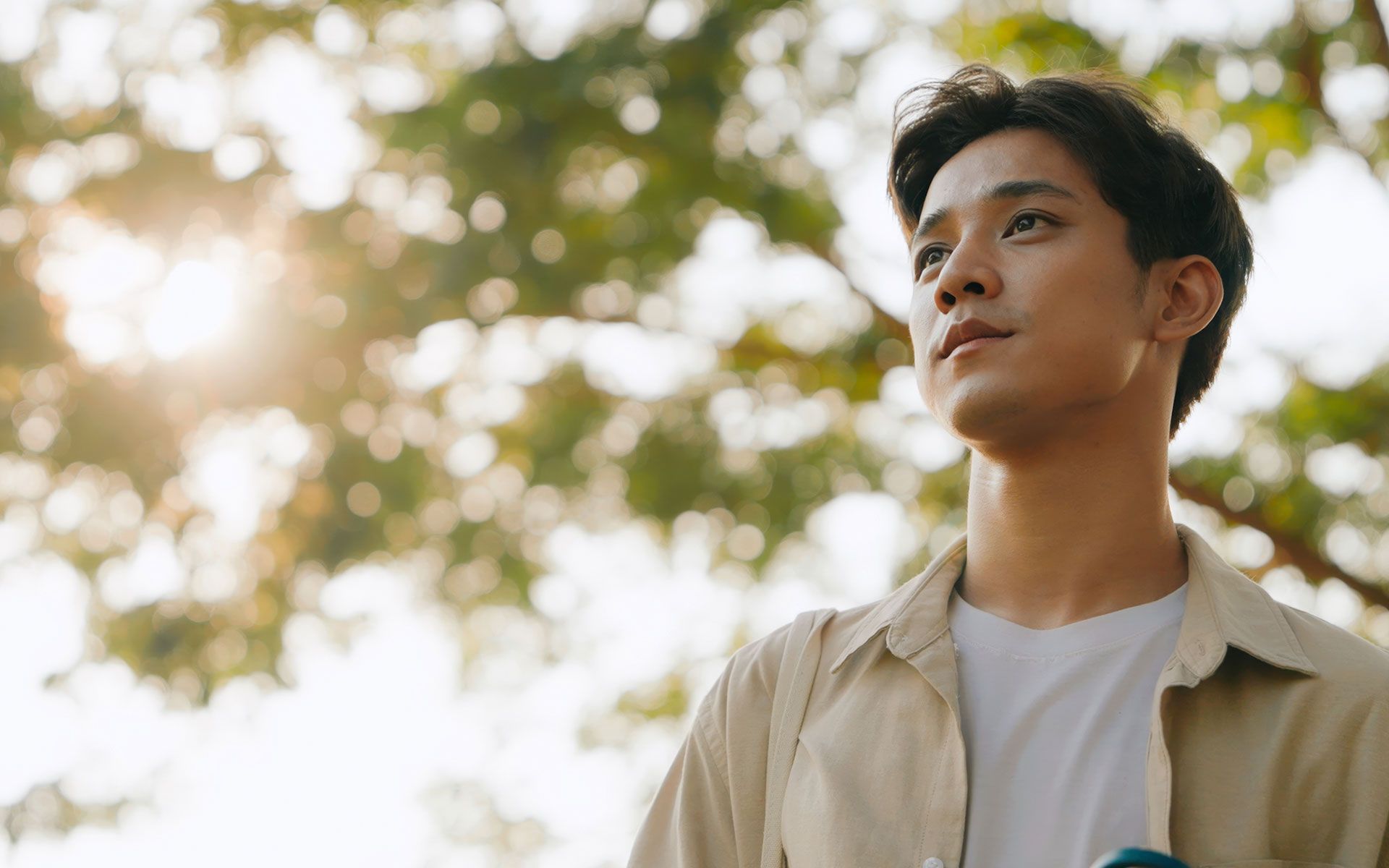 A young man is standing in front of a tree looking up at the sky.