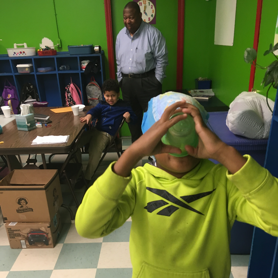 Boy in lime green hoodie looking through green object; two other people in a room.