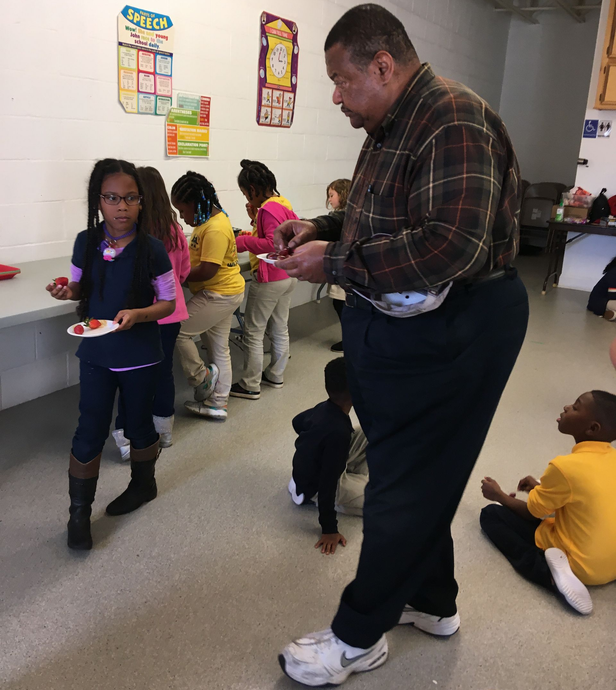 Man serving food to children at a school event. Kids in line, some sitting, with trays.