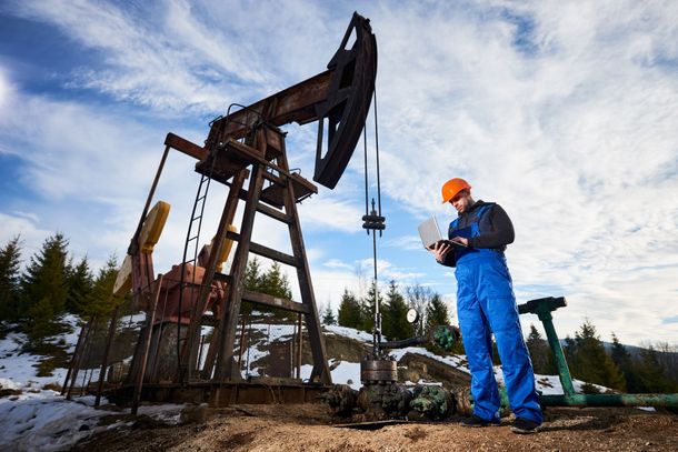 A man is standing in front of an oil pump using a laptop.