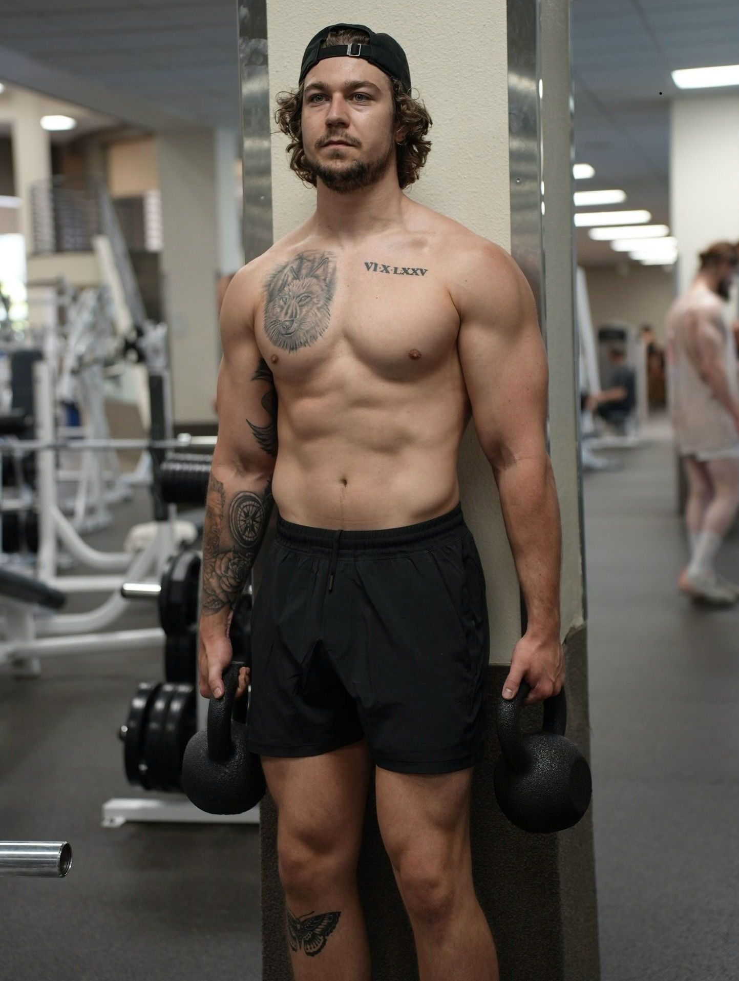 A shirtless man is standing in a gym holding a kettlebell.