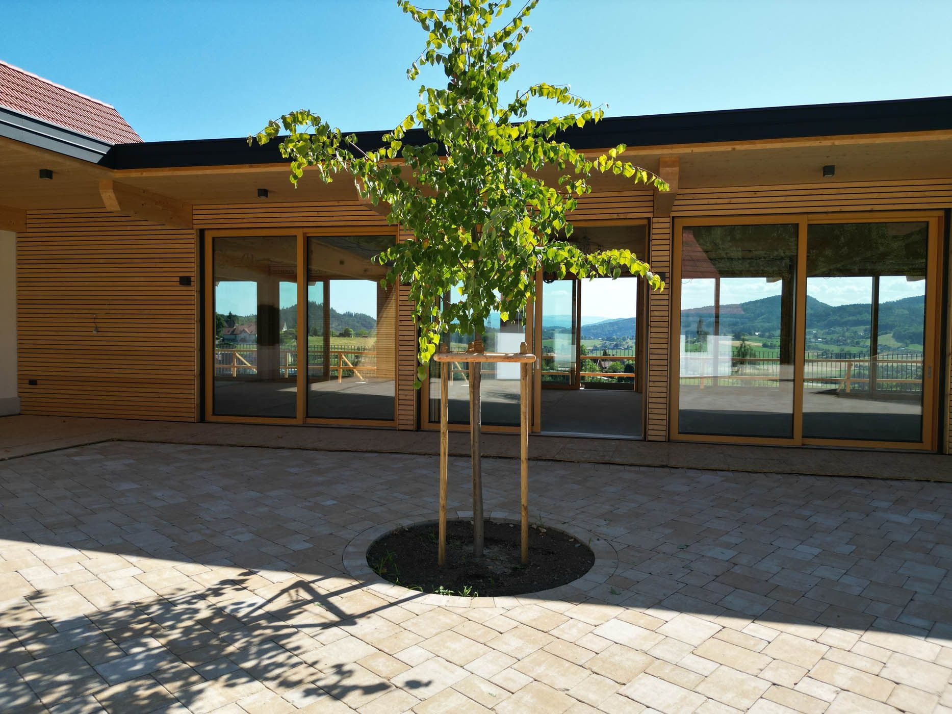 Innenhof mit kleinem Baum, Pflastersteinen und Holzgebäude mit großen Fenstern, im Hintergrund Berge.