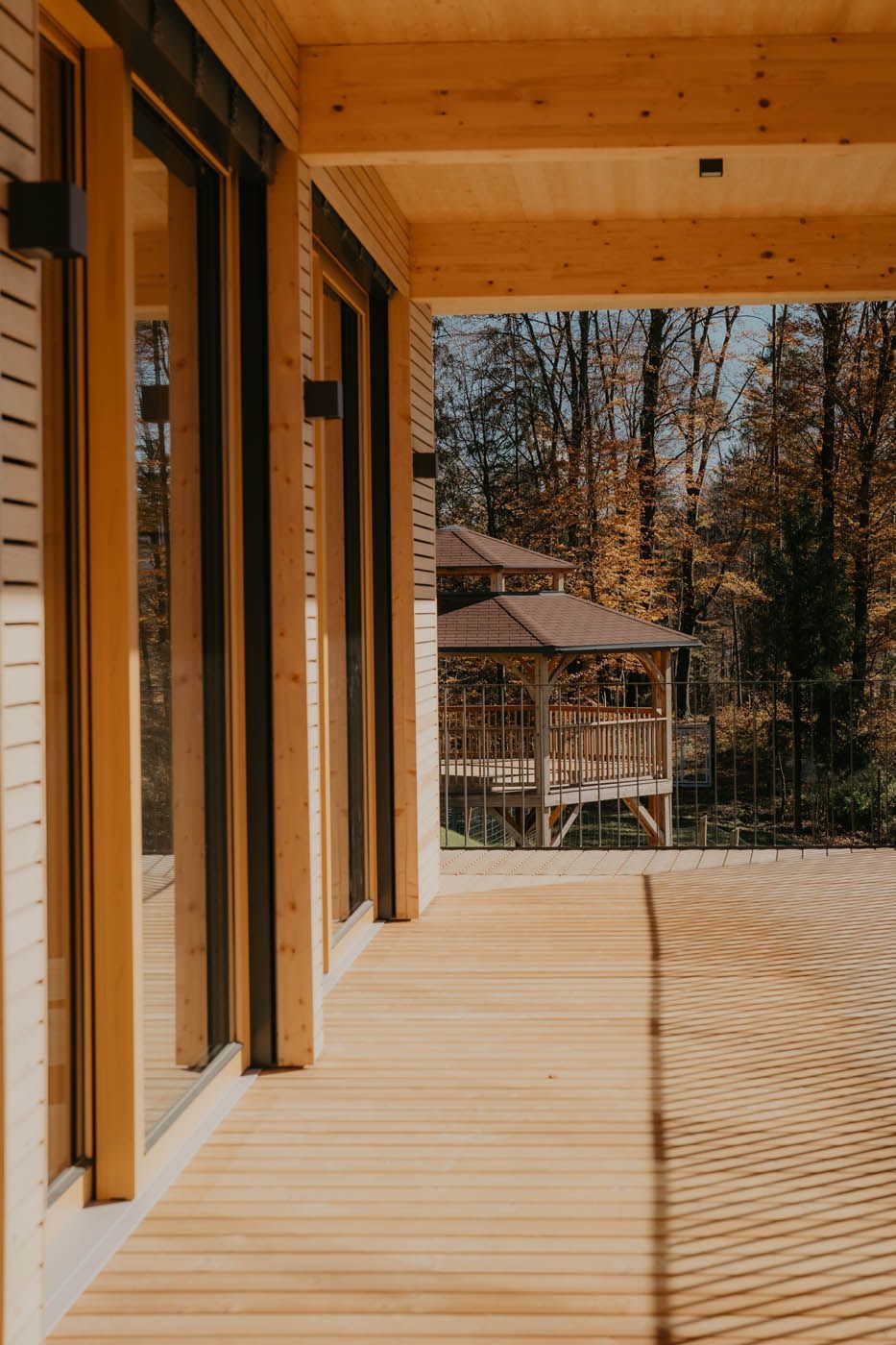 Holzterrasse mit großen Fenstern, die den Blick auf einen Pavillon inmitten eines Waldes freigeben.