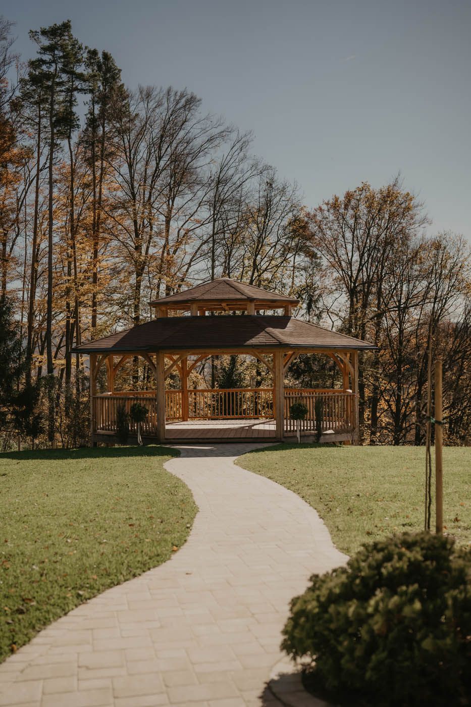 Ein hölzerner Pavillon in einem Park, zu dem ein Weg hinaufführt. An einem sonnigen Tag ist er von Bäumen mit Herbstlaub umgeben.