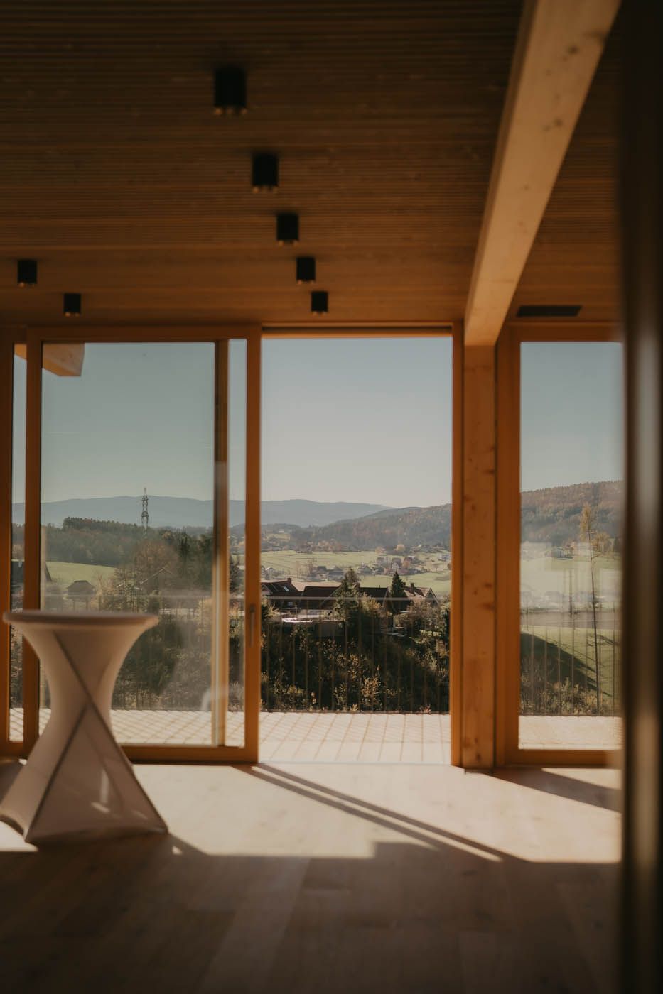 Ein holzgetäfelter Raum mit großen Fenstern, die den Blick auf eine grüne Landschaft und Gebäude in der Ferne freigeben. Im Vordergrund steht ein kleiner Tisch.