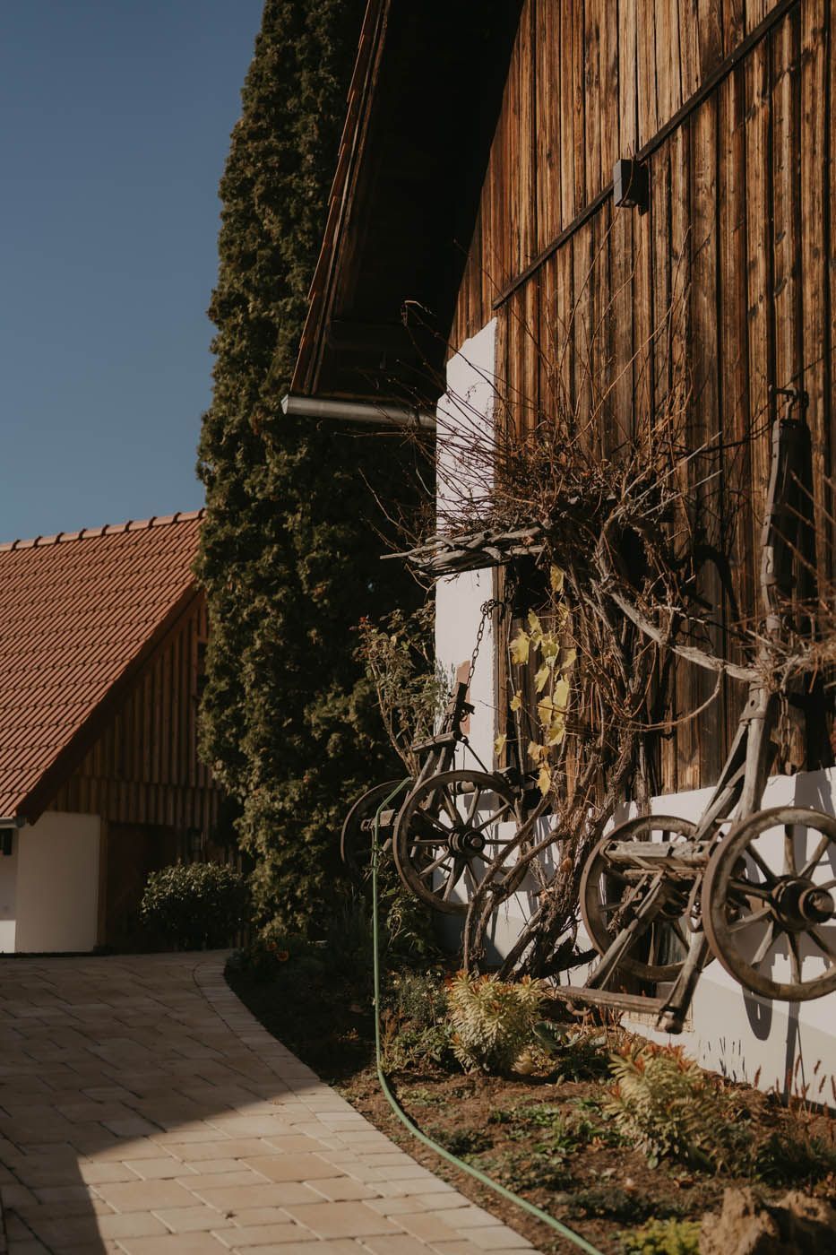 Äußeres Bild einer hölzernen Scheune mit nostalgischer Wagenraddekoration, daneben ein Backsteinweg und hohe grüne Bäume an einem sonnigen Tag.