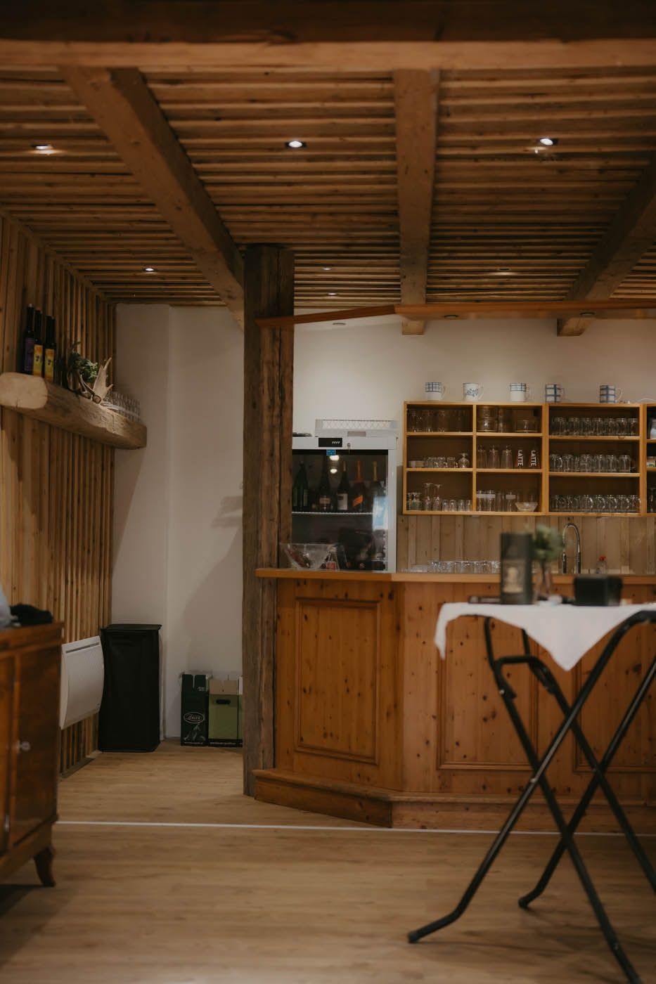 Eine hölzerne Bar in einem rustikalen Raum mit Holzbalken und Regalen voller Gläser. Im Vordergrund steht ein Tisch mit weißem Tischtuch.