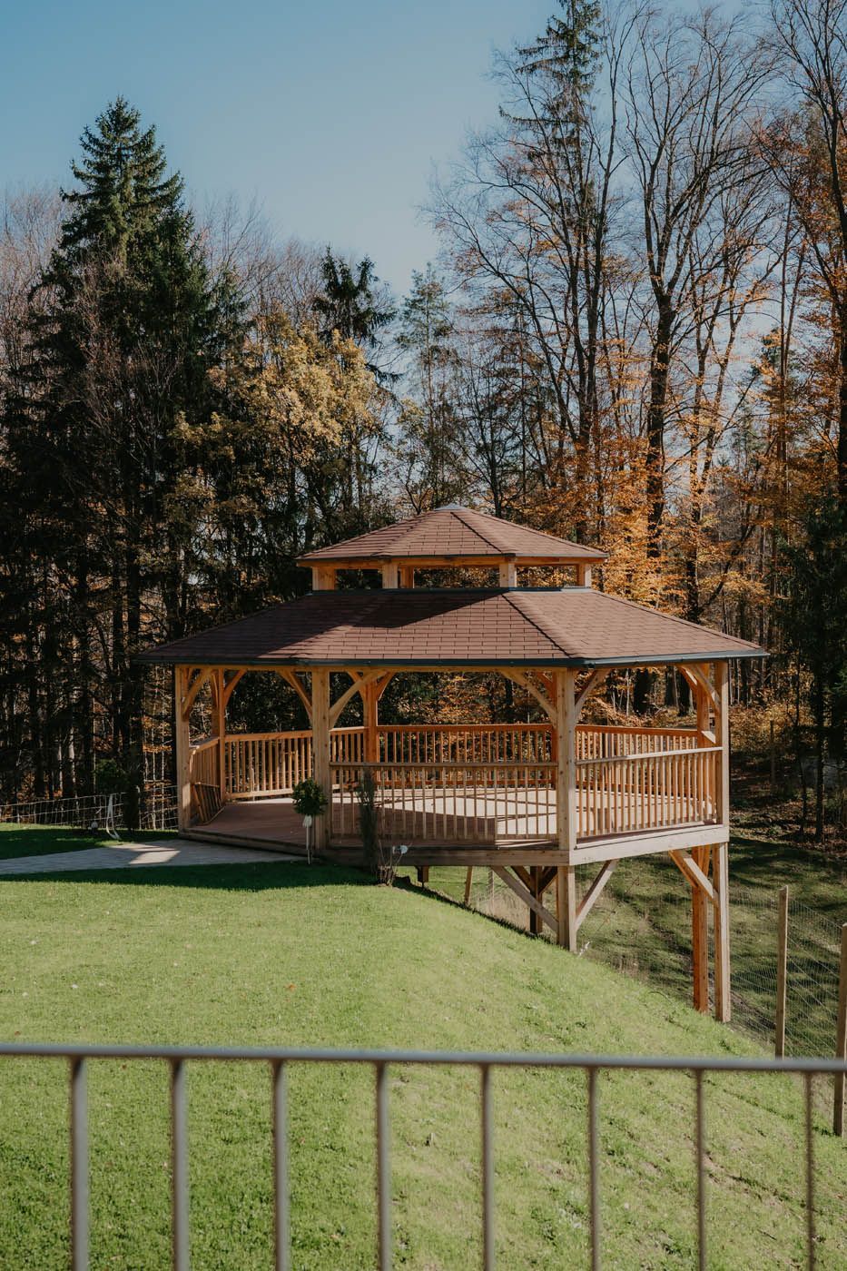 Ein hölzerner Pavillon auf einer Rasenfläche mit Bäumen im Hintergrund. Braune und grüne Farben dominieren das Bild.