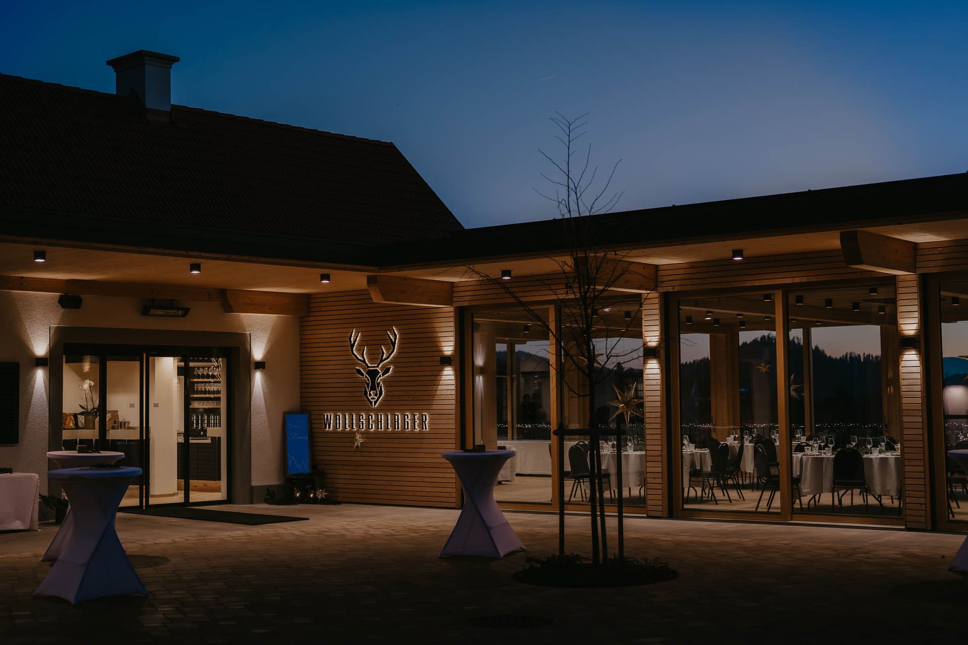 Außenansicht des Restaurants in der Abenddämmerung mit Holzfassade, die von Scheinwerfern beleuchtet wird; der Außenbereich mit Sitzgelegenheiten ist sichtbar.