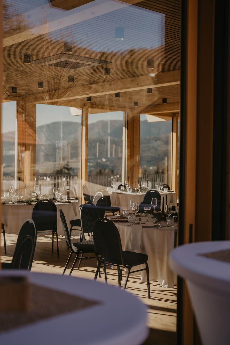 Tische für den Hochzeitsempfang mit weißen Tischdecken und dunklen Stühlen, eingerahmt von einem Fenster mit Bergblick.
