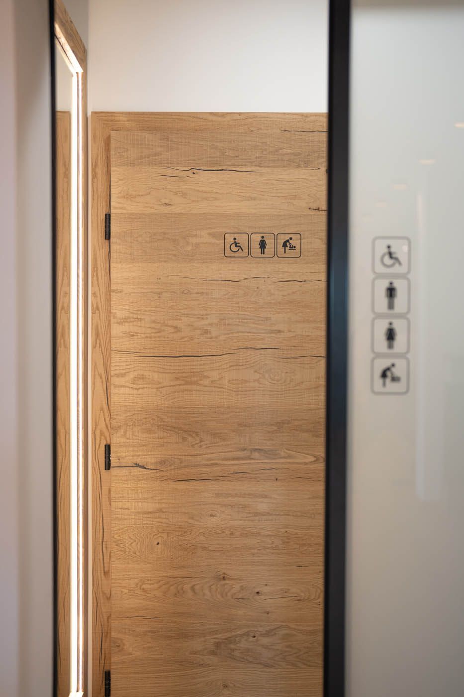 Eine hölzerne Toilettentür mit Symbolen für Barrierefreiheit, daneben eine Glaswand mit ähnlichen Symbolen.