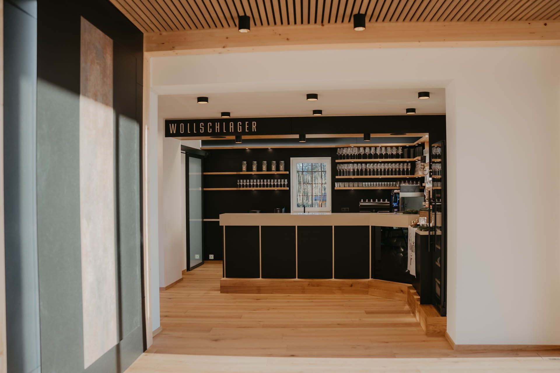 Innenansicht einer Bar mit Holzelementen, schwarzer Theke, Regalen und einem Schild mit der Aufschrift „WEISSBIER“.