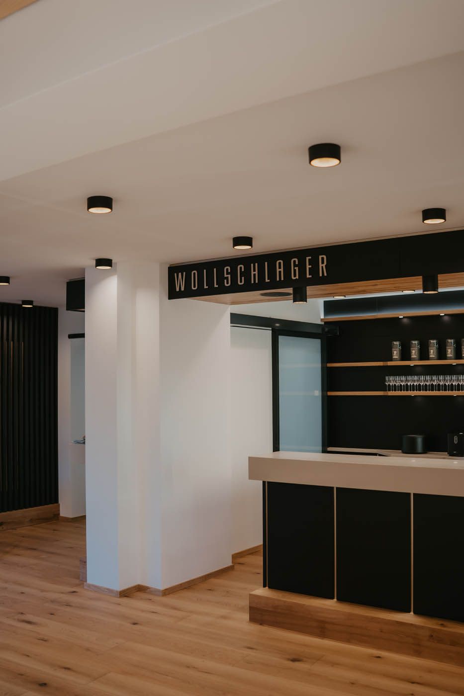 Interieur mit Bar und Schild „NDILSCHLÄGER“. Schwarze Akzente, Holzboden und Deckenleuchten.