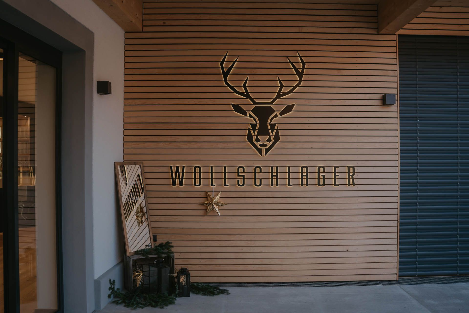 Holzwand mit goldenem Hirschkopf-Logo und dem Schriftzug „WOILSCHIAGER“; Außeneingang.