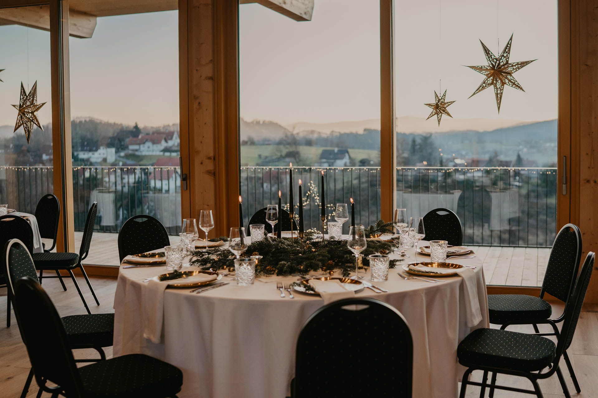 Elegant gedeckter runder Tisch für ein Abendessen, mit einem Kranz als Mittelstück, Kerzen und einem malerischen Ausblick durch ein großes Fenster.