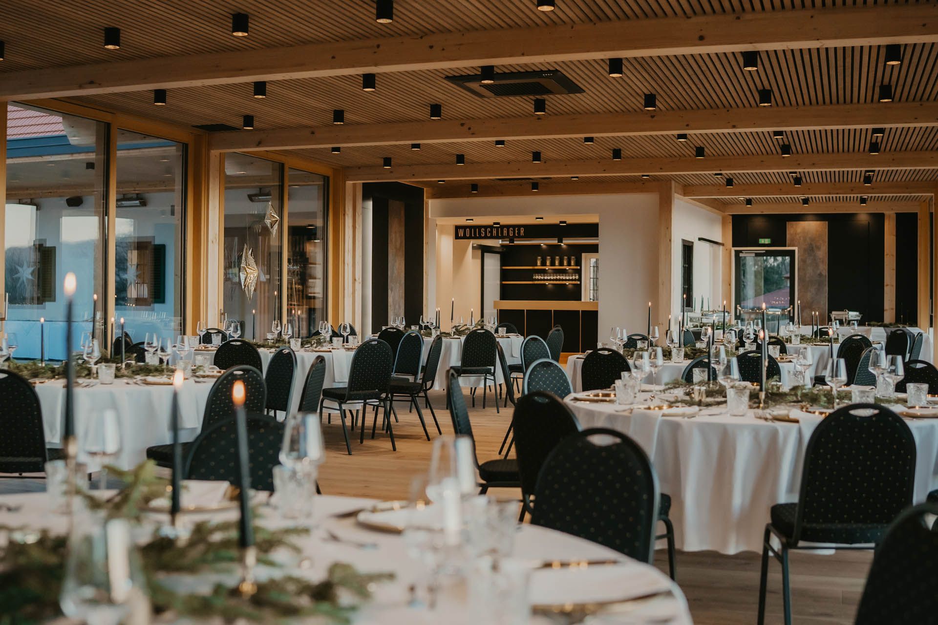 Eleganter Veranstaltungsraum mit runden Tischen, die für ein geselliges Beisammensein gedeckt sind, ausgestattet mit schwarzen Stühlen, Kerzen und Grünpflanzen.