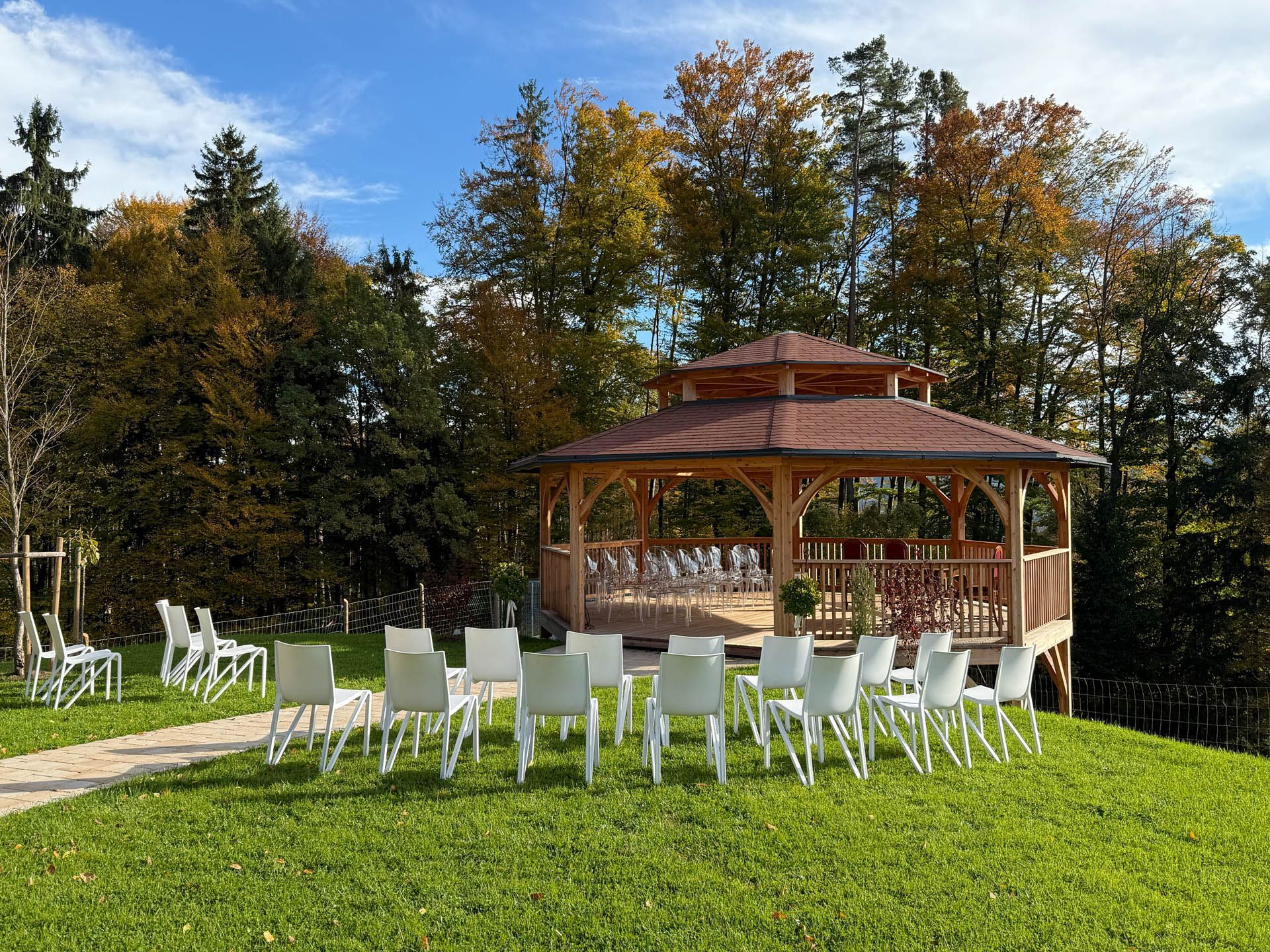 Ein hölzerner Pavillon mit weißen Stühlen auf einer grünen Rasenfläche, im Hintergrund Herbstbäume unter blauem Himmel.