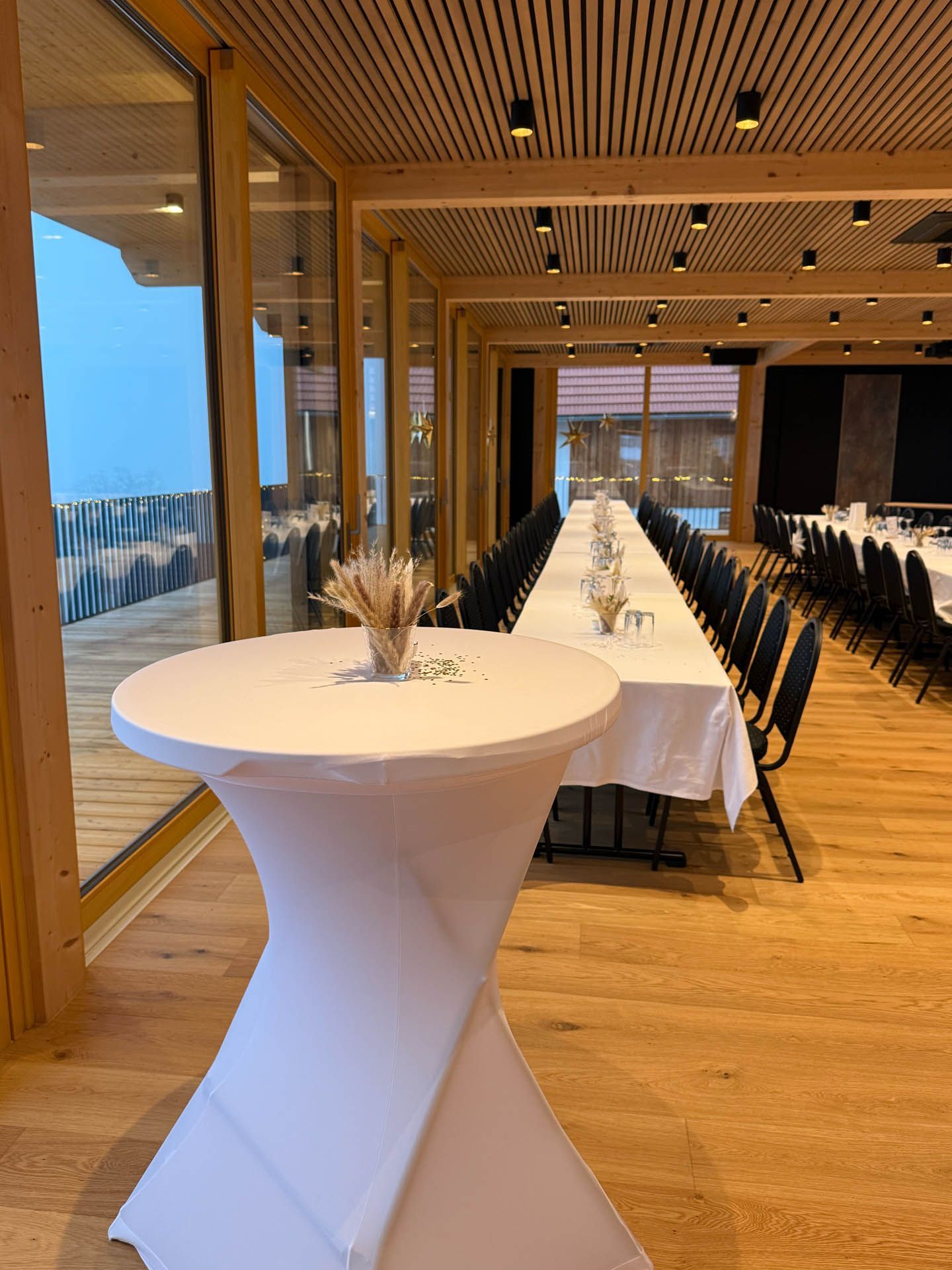 Veranstaltungsraum mit rundem Stehtisch und langen, für Mahlzeiten gedeckten Tischen. Holzelemente und große Fenster.