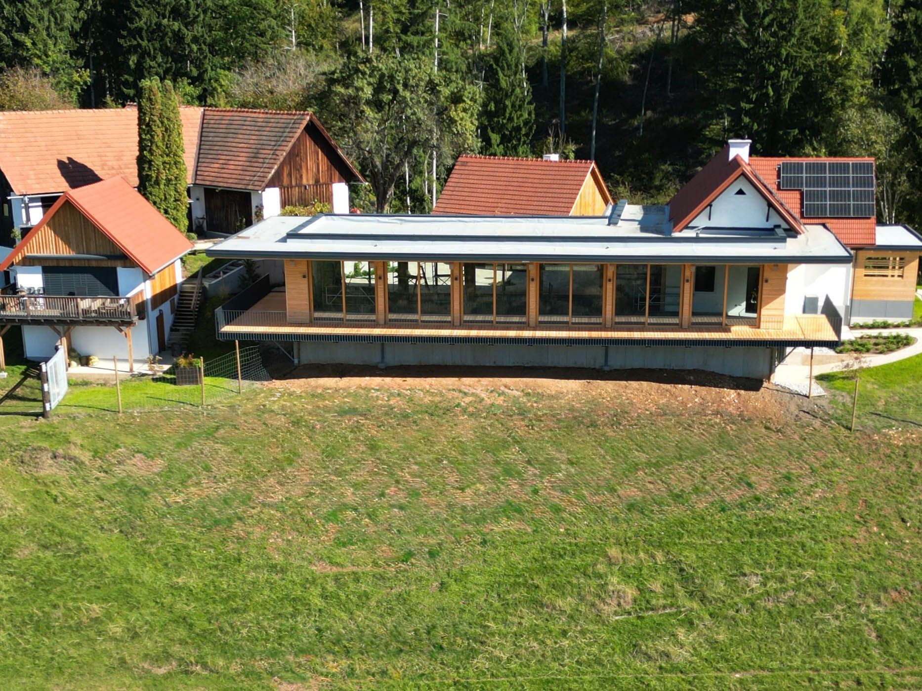 Modernes, langgestrecktes Haus mit großen Fenstern und Holzterrasse, eingebettet in eine grüne Hügellandschaft mit Bäumen.