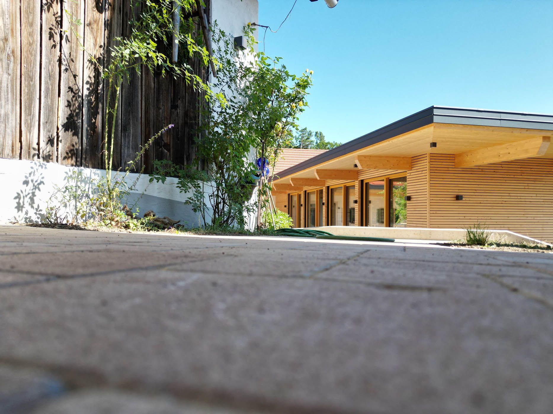 Ansicht aus der Froschperspektive auf ein holzgetäfeltes Gebäude mit mehreren Glastüren und einem gepflasterten Gehweg unter blauem Himmel.