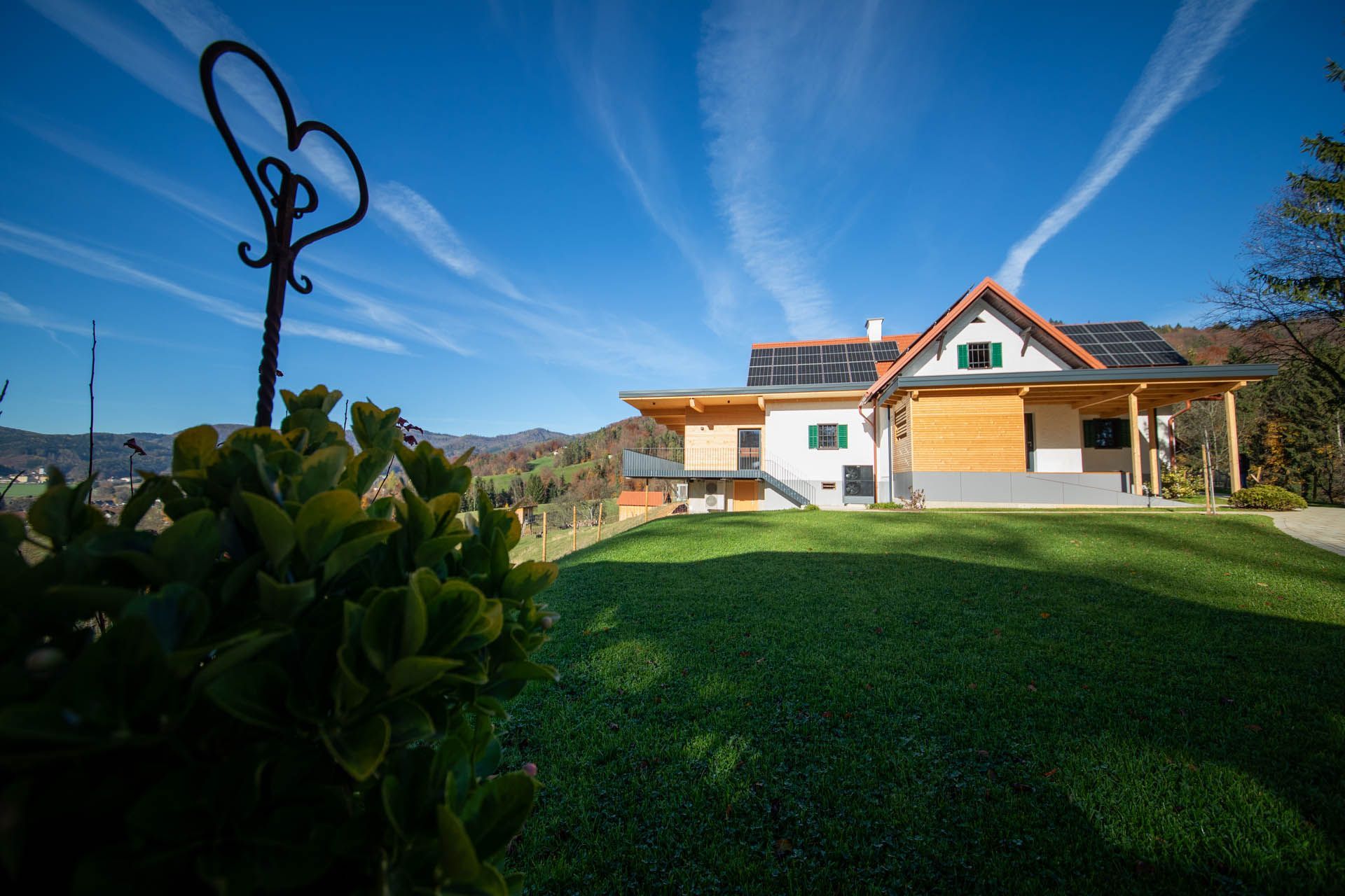 Haus mit Solarpaneelen auf einem grünen Rasen vor blauem Himmel mit Kondensstreifen und einer herzförmigen Metallskulptur.