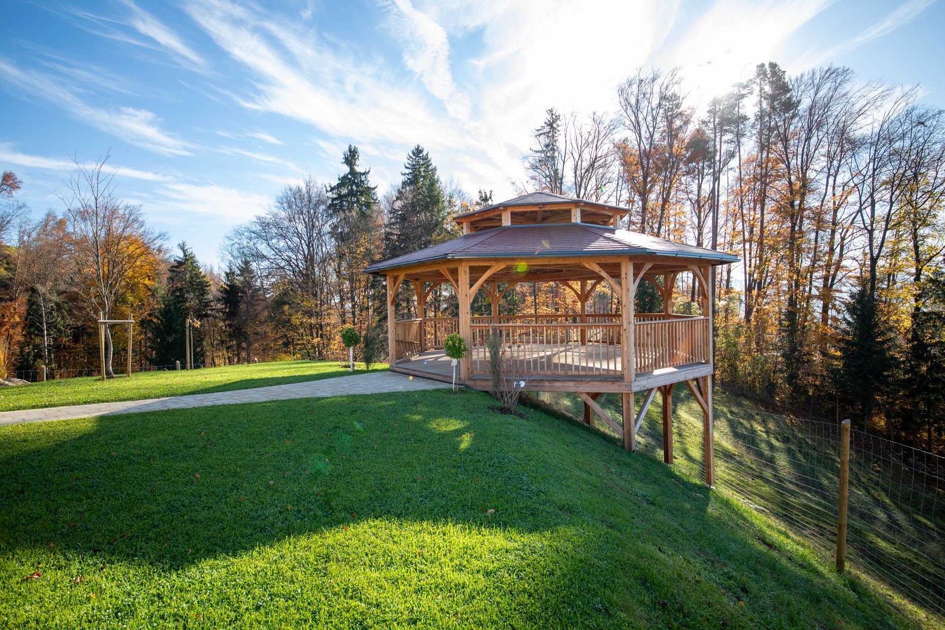 Ein hölzerner Pavillon auf einem grasbewachsenen Hügel mit Blick auf einen Wald unter blauem Himmel.