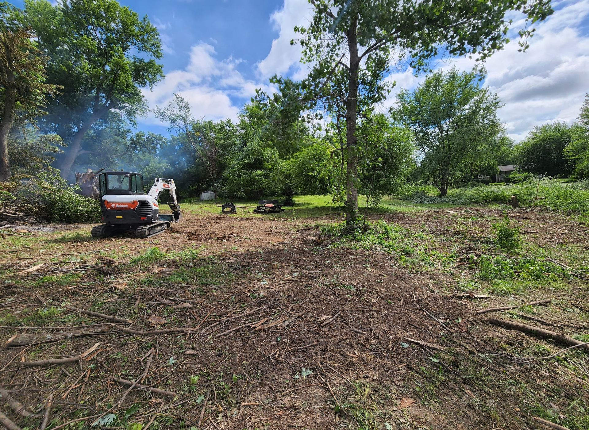 Bobcat clearing brush in a sunny field surrounded by trees. Blue sky, earth-toned ground, white machine.