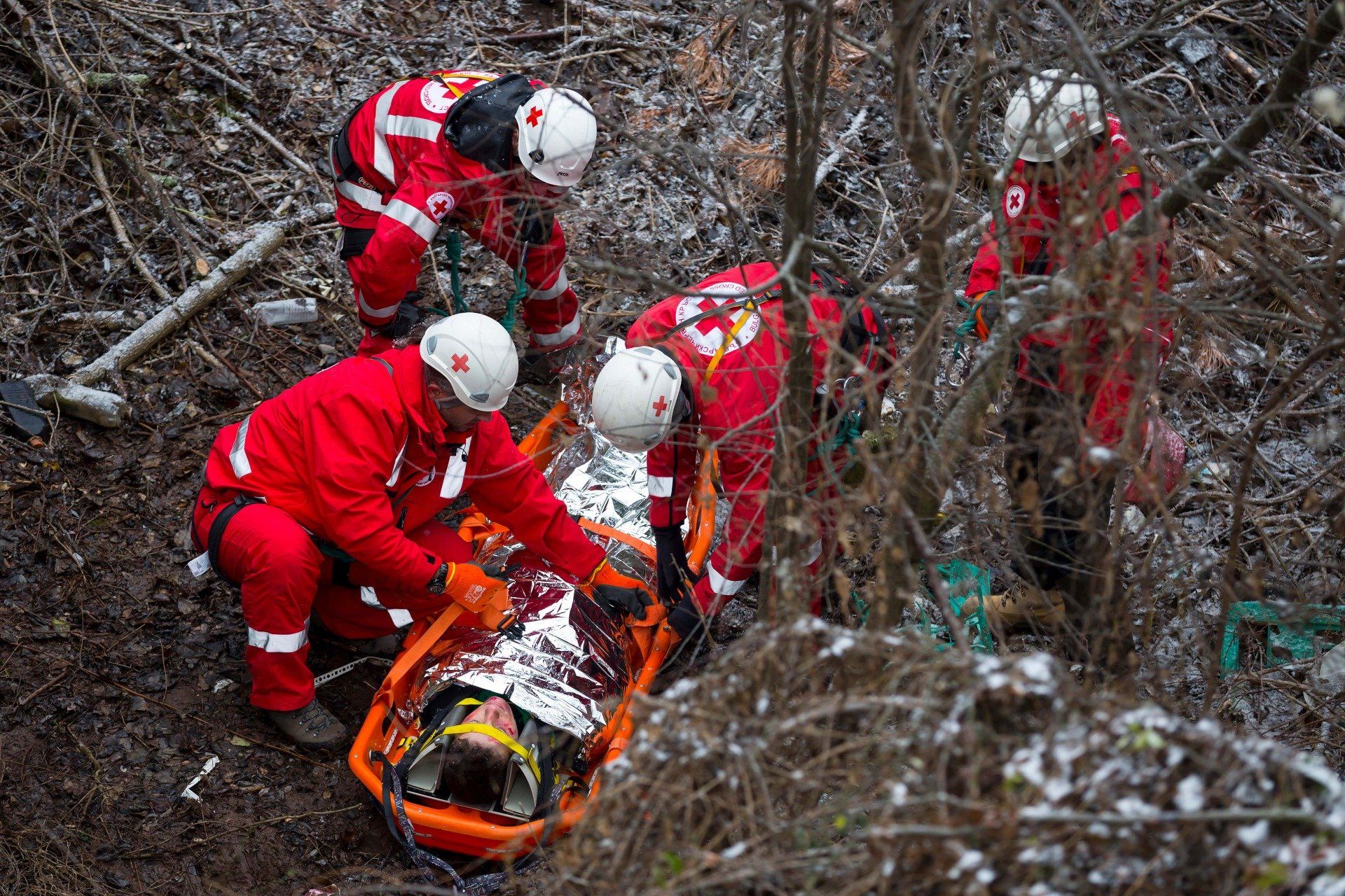Red Cross personnel attending to a person on a stretcher. They are in a wooded area, likely rescuing someone.