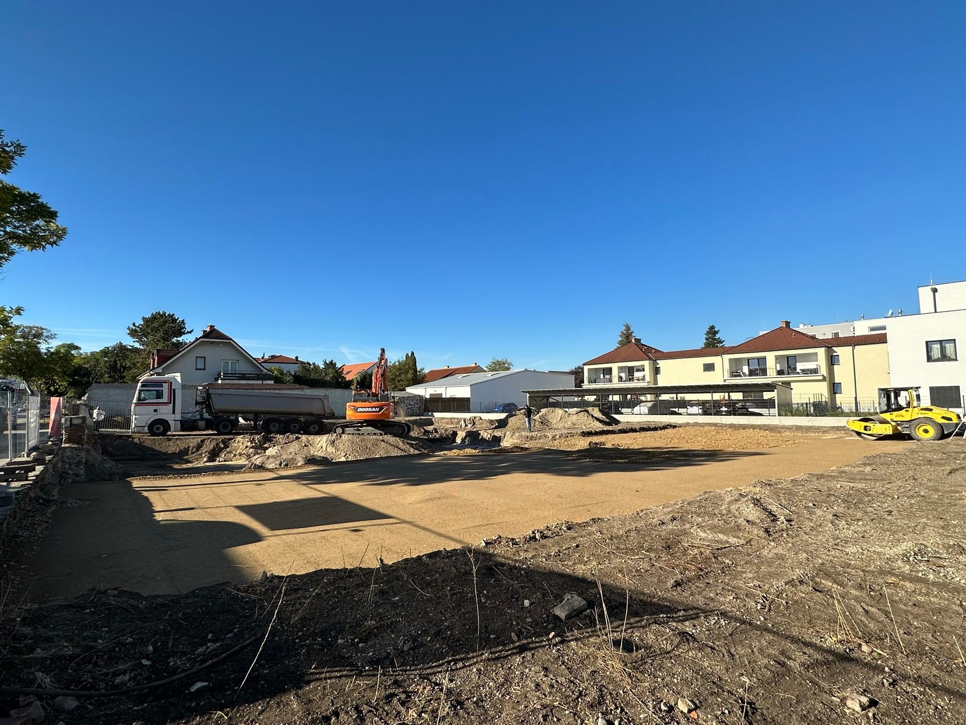 Baustelle mit Bagger, Muldenkipper und ebenem Boden, im Hintergrund Gebäude und blauer Himmel.