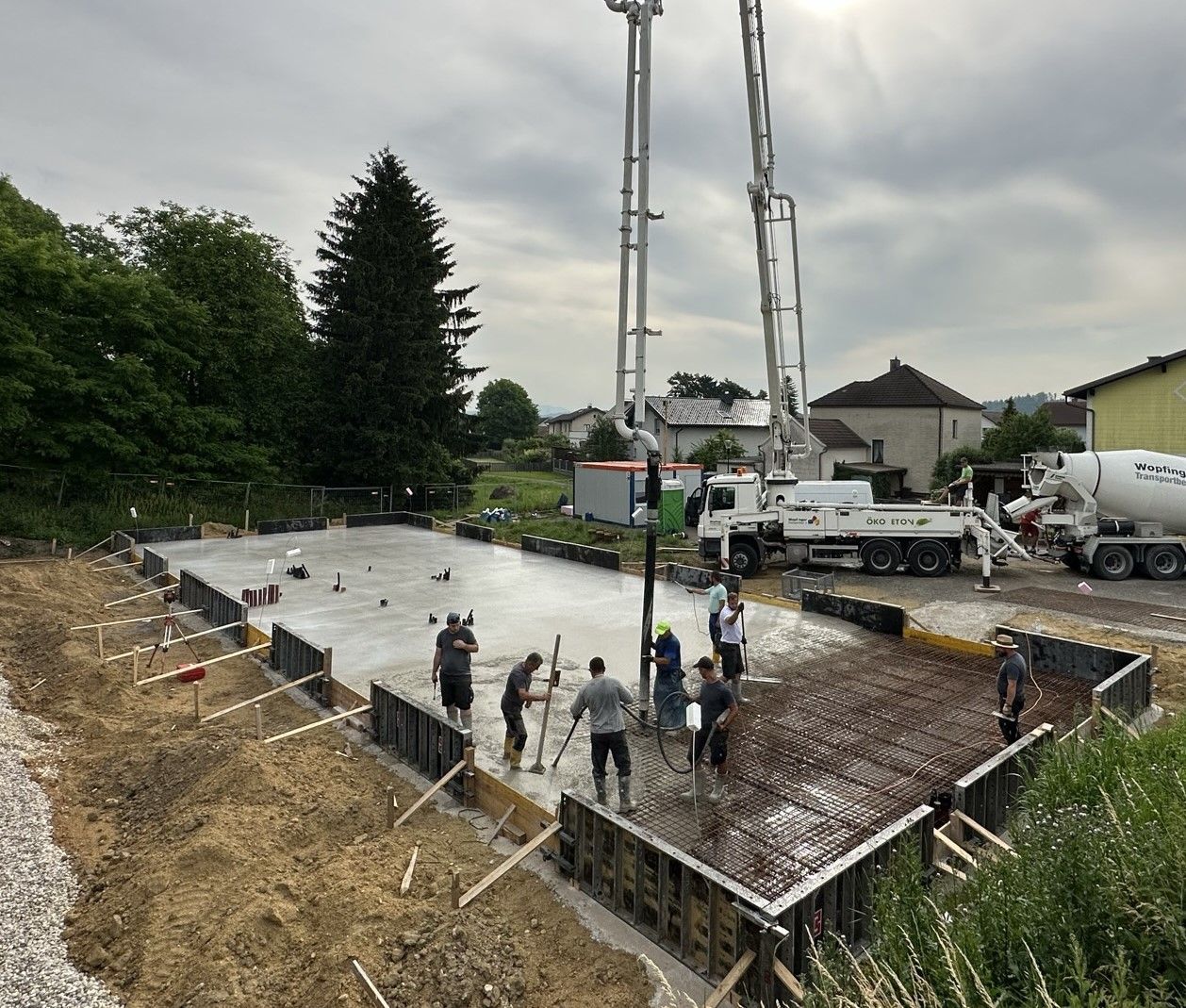 Bauarbeiter gießen Betonfundament. Lastwagen, Pumpe, Bewehrungsstahl und Schalung sind im Freien sichtbar.