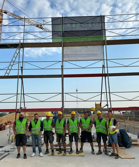 Eine Gruppe von Bauarbeitern in Westen und Schutzhelmen steht unter klarem Himmel vor einem Gerüst.