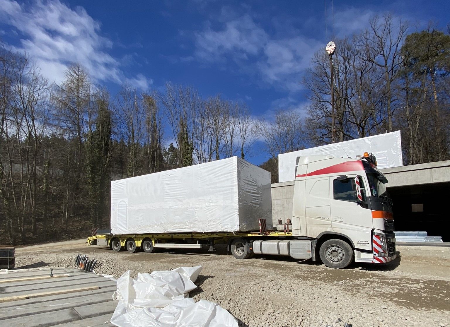 LKW mit großer, weißer, rechteckiger Ladung auf einem Pritschenanhänger in einer Baustelle. 