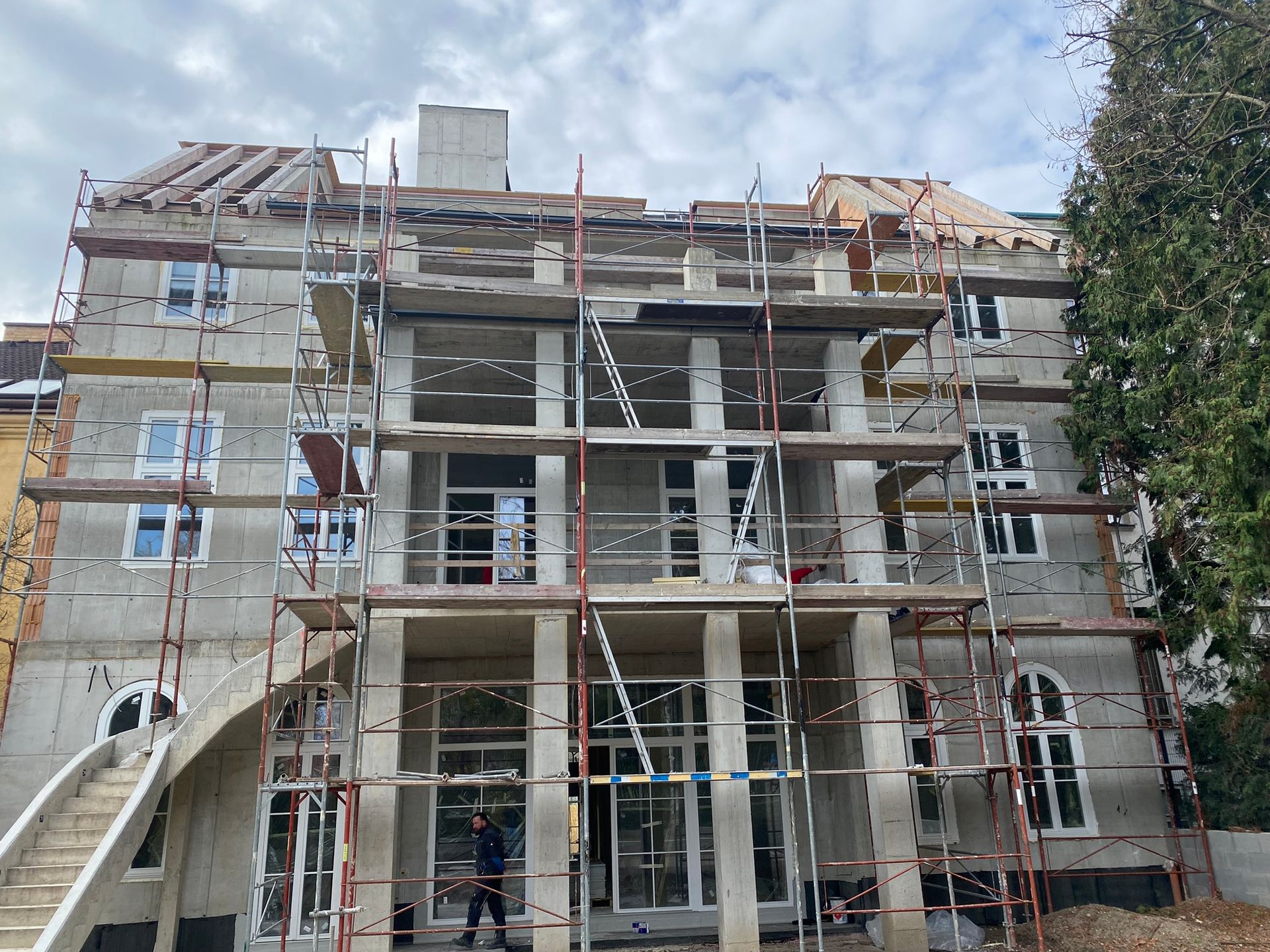 Gebäude im Bau mit Gerüst; grauer Beton, weiße Fenster und hölzerne Dachstützen.