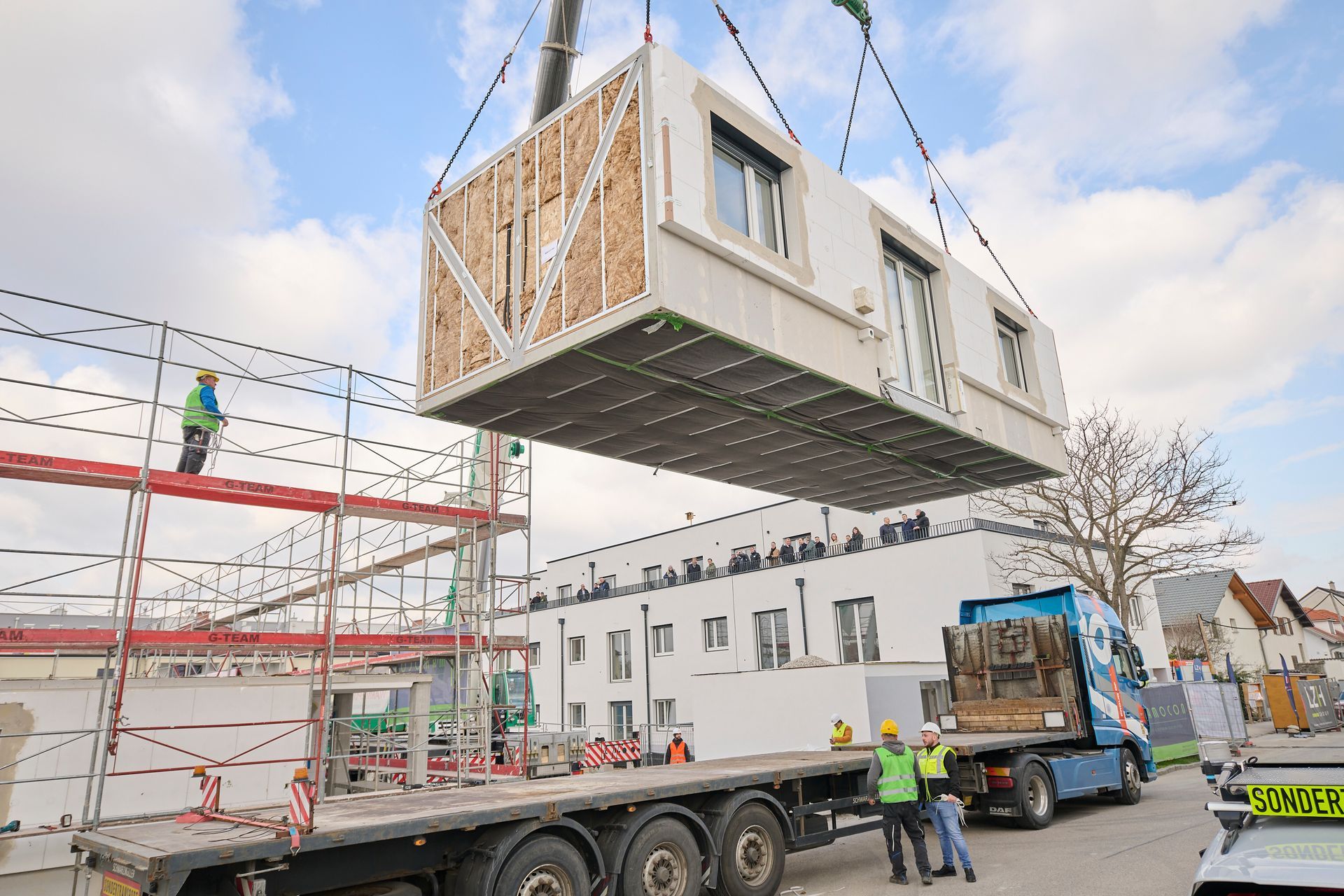 Ein Kran hebt eine modulare Baueinheit an, Arbeiter auf einer Baustelle mit einem LKW schauen zu.