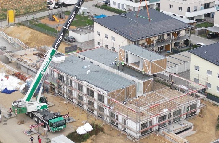 Ein Kran hebt einen modularen Gebäudeabschnitt auf eine mehrstöckige Baustelle.