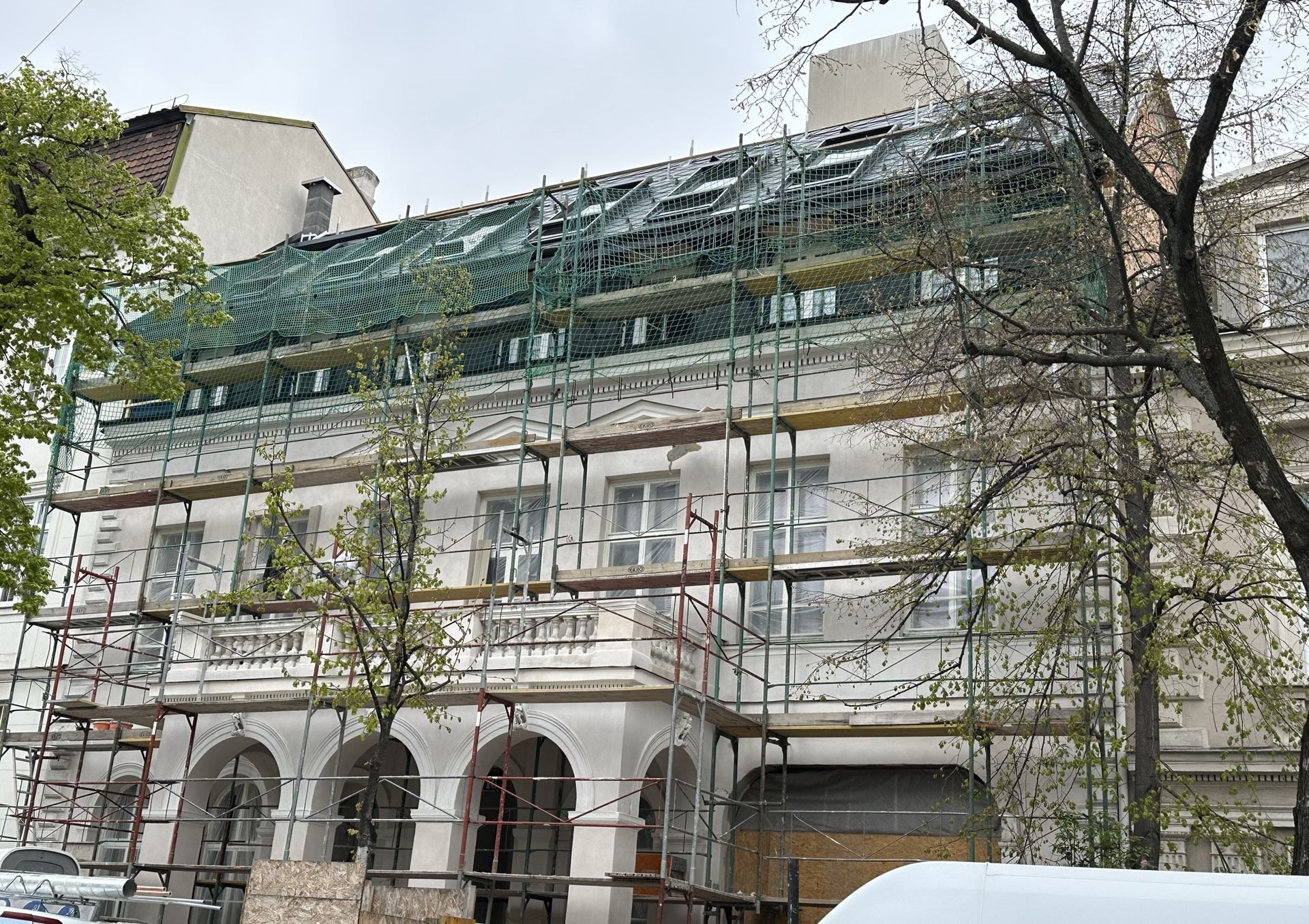 Gebäude wird mit Gerüst und Netzen einer Dachreparatur unterzogen. Weiße Fassade, sichtbare Fenster, bewölkter Himmel.