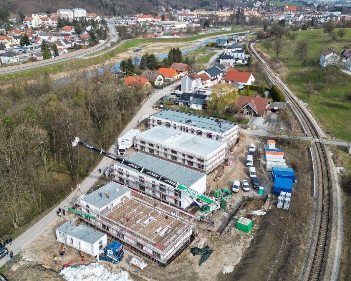 Baustelle mit modularen Gebäuden in der Nähe einer Eisenbahnlinie und eines Wohngebiets. Kran, Arbeiter und Materialien.
