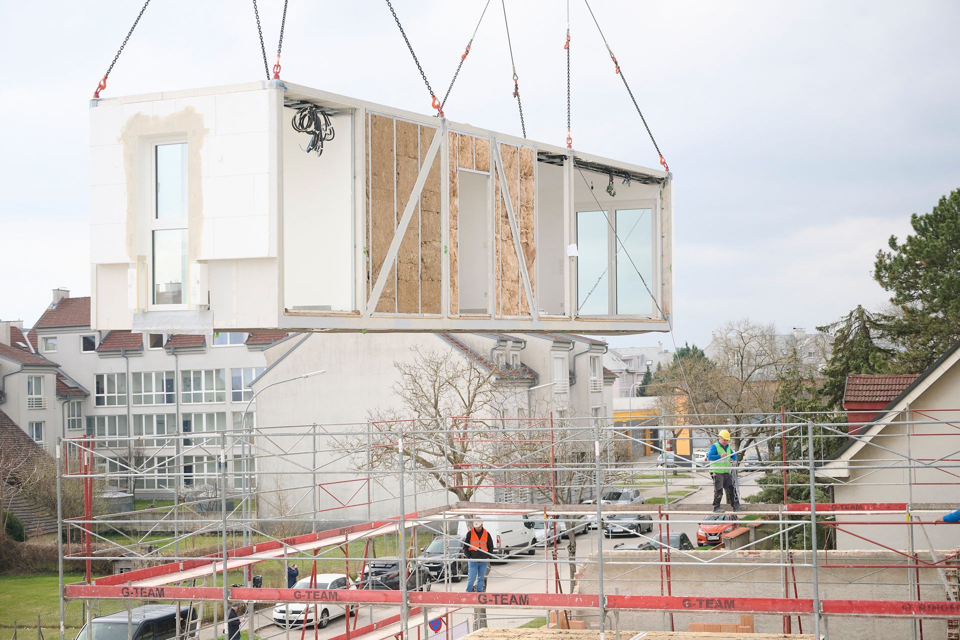 Ein Fertighaus wird mit einem Kran angehoben, darunter stehen Arbeiter auf einem Gerüst.