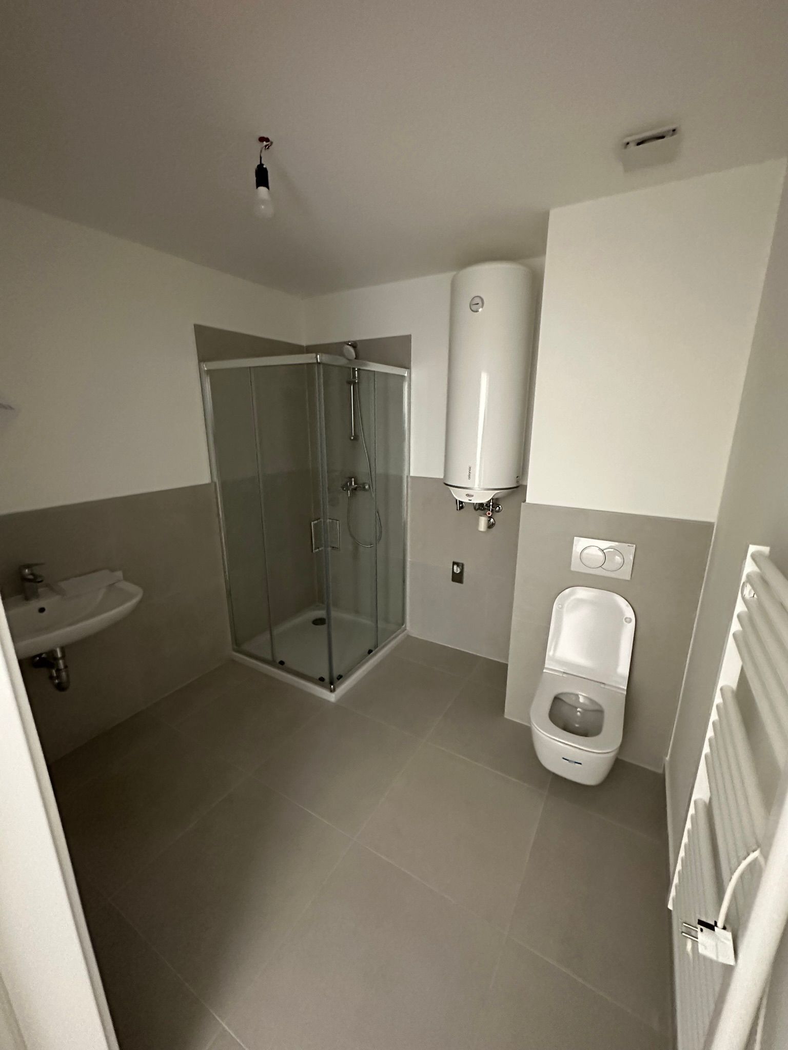 Badezimmer mit grau gefliestem Boden, Dusche, Toilette, Waschbecken und Warmwasserbereiter.