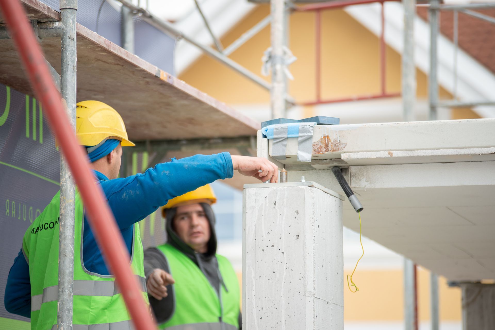 Zwei Bauarbeiter mit gelben Helmen zeigen auf den Stützbalken eines Gebäudes.
